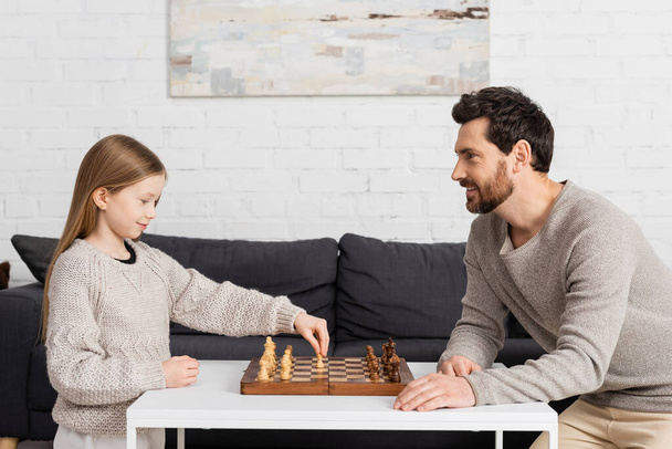 vista laterale della ragazza intelligente che gioca a scacchi con il padre allegro in soggiorno  - Foto, immagini
