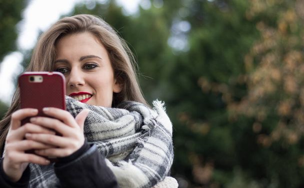Close up de menina loira segurando telefone celular vermelho no dia de outono frio no parque. Mulher jovem, vício em celular, aplicativo de namoro on-line, comunicação, novas tecnologias, conceitos da moda - Foto, Imagem