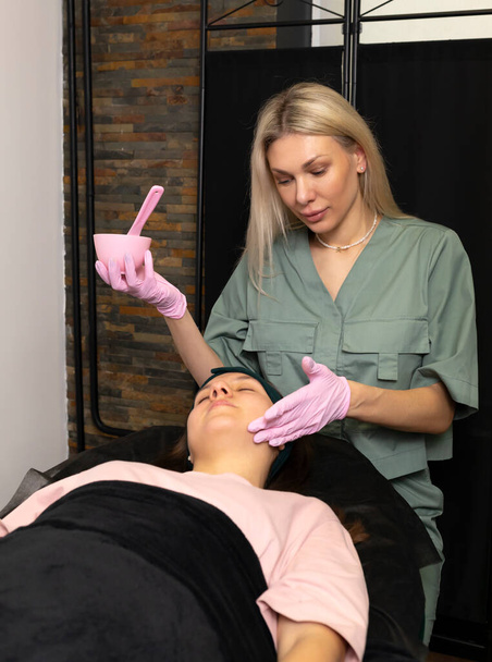 Acne treatment, young skin care. Cosmetologist does alginate mask procedure on female teen face. Teenage girl lying on medical couch. Dermatologist in pink gloves holds bowl, brush. Vertical plane. - Фото, зображення