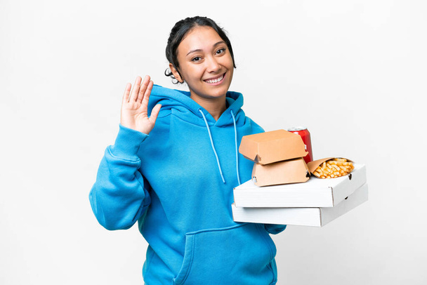 Young Uruguayan woman holding pizzas and burgers over isolated white background saluting with hand with happy expression - Фото, изображение