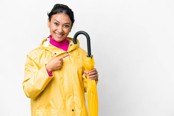 Young Uruguayan woman with rainproof coat and umbrella over isolated white background pointing to the side to present a product - Φωτογραφία, εικόνα