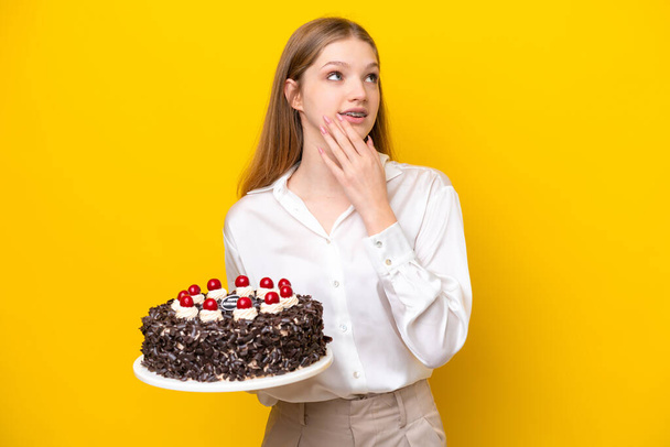 Teenager rusky dívka drží narozeninový dort izolované na žlutém pozadí při pohledu nahoru s úsměvem - Fotografie, Obrázek