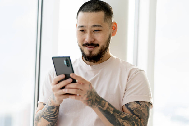 Smiling Asian man with stylish tattoos holding  smartphone using mobile app shopping online at home. Modern bearded hipster reading text message, ordering food, communication. Technology concept - Fotó, kép