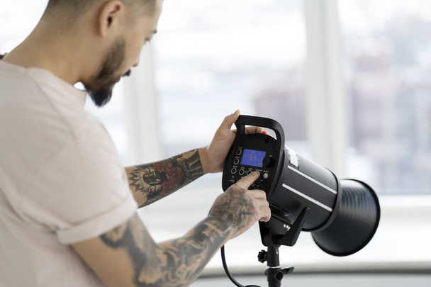 Professional asian photographer with stylish tattoos setting up lighting in modern studio. Assistant preparing equipment for photo shoot, selective focus  - Photo, Image