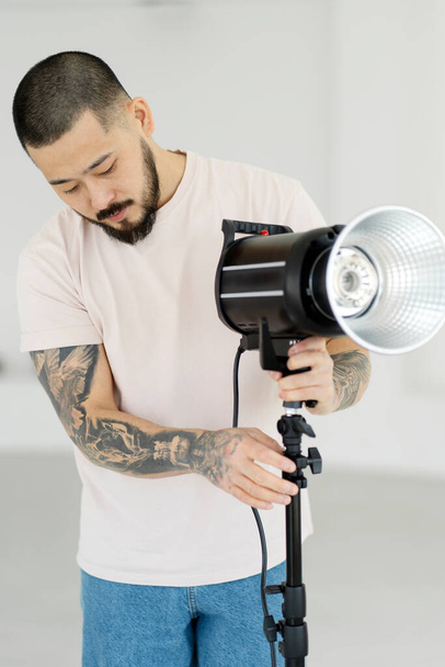 Portret van professionele Aziatische fotograaf met stijlvolle tattoos die verlichting opzetten in een moderne studio. Assistent voorbereiding voor fotoshoot - Foto, afbeelding
