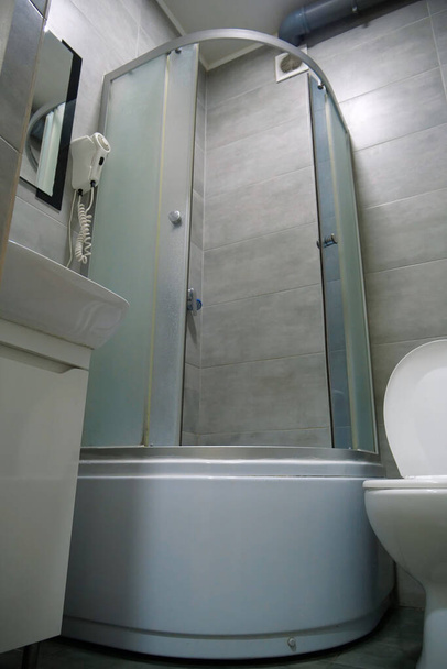 Δωμάτιο τουαλέτας εσωτερικό με λευκή λεκάνη τουαλέτας με ανοιχτό καπάκι, καμπίνα ντους, νιπτήρα, στεγνωτήρα μαλλιών, ντουλάπι κάτω από το νιπτήρα και γκρι πλακάκια τοίχου και δαπέδου - Φωτογραφία, εικόνα