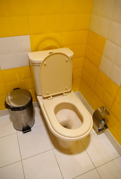 Δωμάτιο τουαλέτας εσωτερικό με λευκό μπωλ τουαλέτας με ανοιχτό καπάκι, μεταλλικό καλάθι απορριμμάτων, βούρτσα τουαλέτας και κίτρινα πλακάκια τοίχου - Φωτογραφία, εικόνα