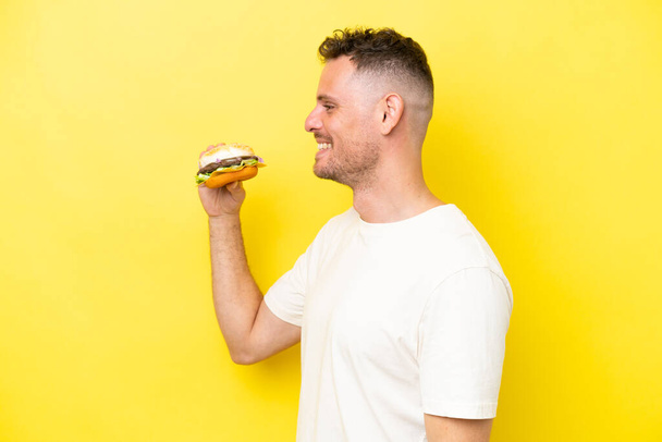 Young caucasian man holding a burger isolated on yellow background laughing in lateral position - Photo, Image