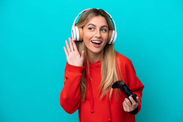 Young Uruguayan woman playing with a video game controller isolated on blue listening to something by putting hand on the ear - Foto, immagini