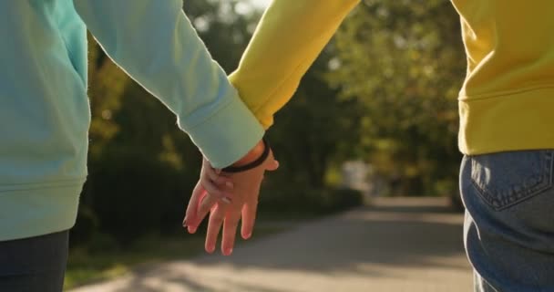 Girl friends hold hands walking in park illuminated by bright sunlight. Teenagers in blue and yellow sweaters demonstrate strong friendship - Imágenes, Vídeo