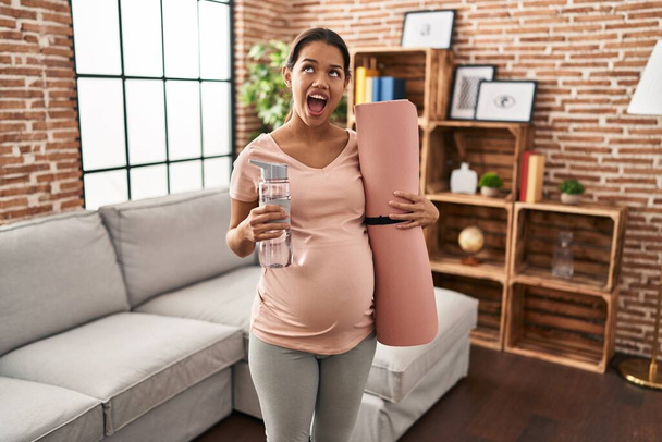 Νεαρή έγκυος γυναίκα κρατώντας στρώμα γιόγκα στο σπίτι θυμωμένη και θυμωμένη ουρλιάζοντας απογοητευμένη και έξαλλη, φωνάζοντας με θυμό κοιτάζοντας ψηλά.  - Φωτογραφία, εικόνα