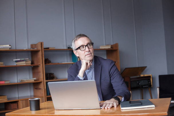 Portrait d'un homme d'affaires adulte en lunettes et costume assis et regardant un ordinateur portable sur le fond du mur de bureau - Photo, image