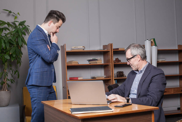 Зрелый серьезный бизнесмен в темно-синем костюме со своим партнером стоят за ноутбуком в офисе и думают о проектах - Фото, изображение