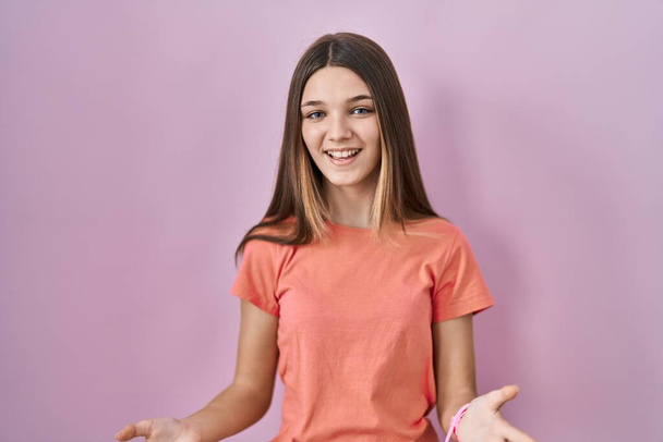 Teenager girl standing over pink background smiling cheerful with open arms as friendly welcome, positive and confident greetings  - 写真・画像