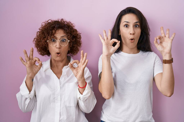 Ισπανόφωνη μητέρα και κόρη φορώντας casual λευκό t πουκάμισο πάνω από ροζ φόντο φαίνεται έκπληκτος και σοκαρισμένος κάνει εντάξει σύμβολο έγκρισης με τα δάχτυλα. τρελή έκφραση  - Φωτογραφία, εικόνα