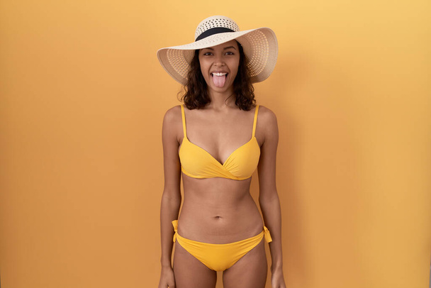 Mulher hispânica jovem usando biquíni e chapéu de verão colando a língua feliz com expressão engraçada. conceito de emoção.  - Foto, Imagem