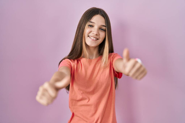 Девушка-подросток, стоящая на розовом фоне и одобряющая позитивный жест рукой, улыбается и радуется успеху. жест победителя.  - Фото, изображение