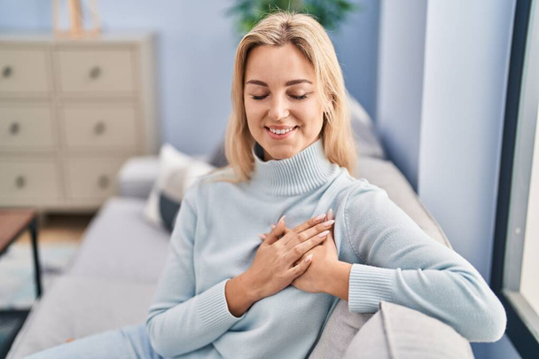 Young blonde woman sitting on sofa with hands on heart at home - Foto, afbeelding