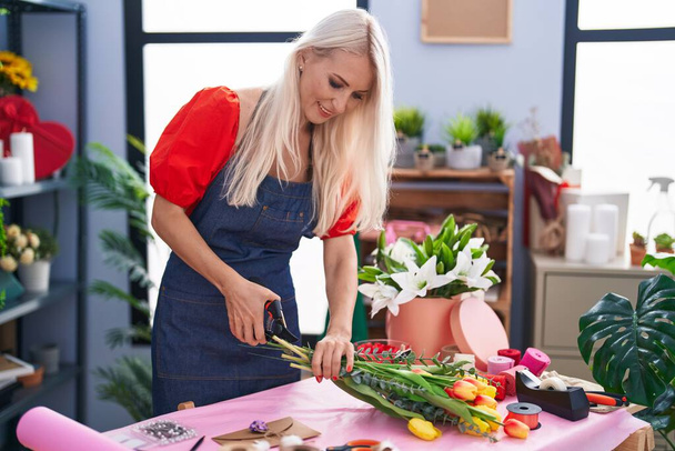 Young blonde woman florist make bouquet of flowers cutting stem at florist store - 写真・画像