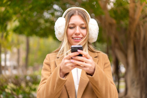 Açık havada kışlık manşet takan güzel, sarışın bir kadın cep telefonuyla mesaj yolluyor. - Fotoğraf, Görsel