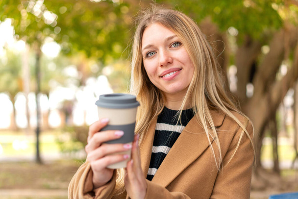 Jeune jolie femme blonde tenant un café à emporter à l'extérieur avec une expression heureuse - Photo, image