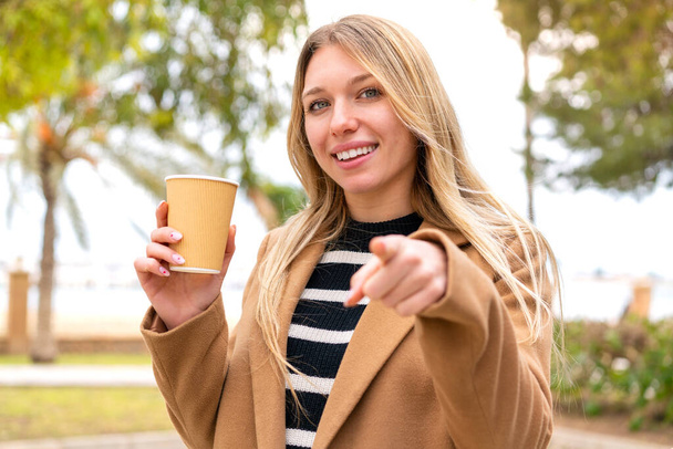 Joven mujer rubia bonita sosteniendo un café para llevar al aire libre señala el dedo en usted con una expresión de confianza - Foto, Imagen