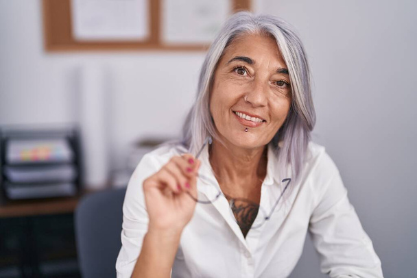 Middle age grey-haired woman business worker smiling confident sitting on table at office - Foto, immagini