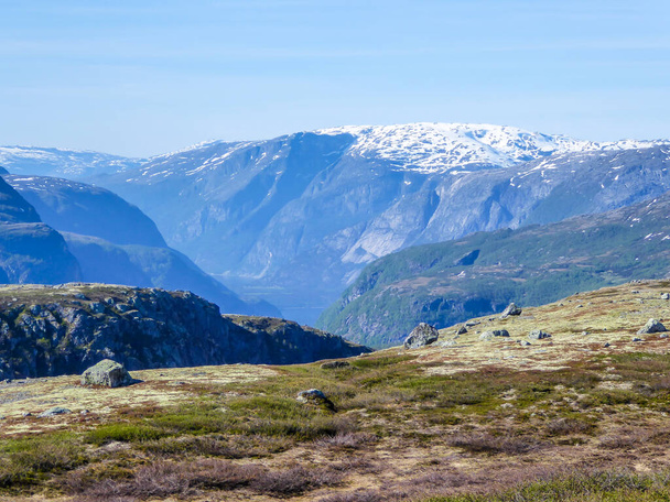 Вид на высшие районы Эйдфьорда, Норвегия. Высокие горы покрыты снегом. Пышная зеленая флора растет на склонах. Небо чистое голубое. В ущелье идет туман из воды. - Фото, изображение