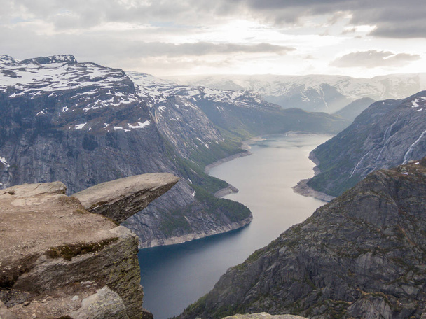 Slavná skalní formace, Trolltunga s výhledem shora na jezero Ringedalsvatnet, Norsko. Skalní. Svahy hor jsou částečně pokryty sněhem. Voda v jezeře je námořně modrá. - Fotografie, Obrázek