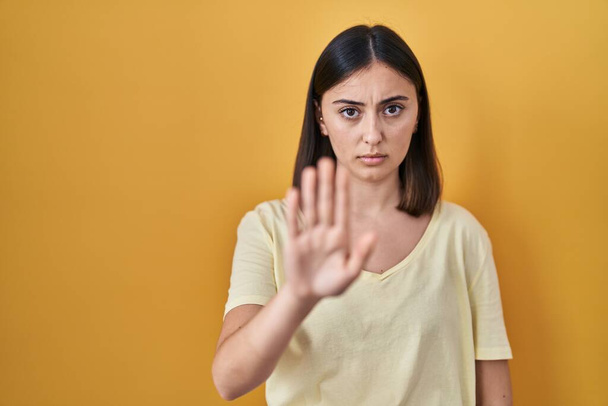 Spaanse meisje dragen casual t-shirt over gele achtergrond doen stoppen met zingen met palm van de hand. waarschuwingsuitdrukking met negatief en ernstig gebaar op het gezicht.  - Foto, afbeelding