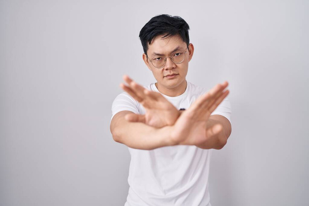 Νεαρός Ασιάτης στέκεται πάνω από λευκό φόντο έκφραση απόρριψης διασχίζει τα χέρια και τις παλάμες κάνει αρνητικό σημάδι, θυμωμένος πρόσωπο  - Φωτογραφία, εικόνα