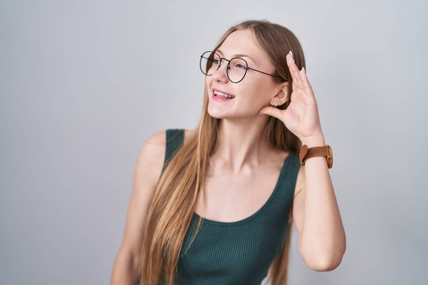 Jeune femme caucasienne debout sur fond blanc souriant avec la main sur l'oreille en écoutant une rumeur ou des commérages. concept de surdité.  - Photo, image