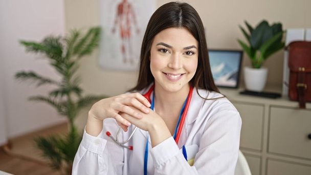 Молода красива іспаномовна жінка лікар посміхається впевнено сидячи на столі в клініці
 - Фото, зображення