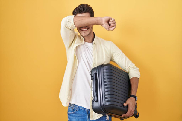 Jonge Spaanse man die een koffer vasthoudt en op zomervakantie gaat glimlachend vrolijk kiekeboe speelt met handen die zijn gezicht laten zien. verrast en verlaten  - Foto, afbeelding