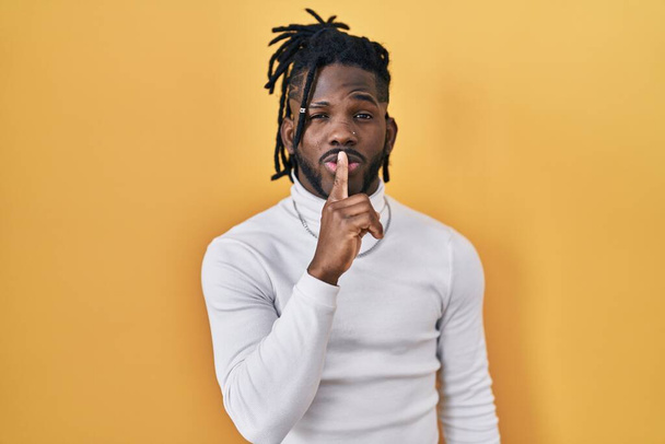 African man with dreadlocks wearing turtleneck sweater over yellow background asking to be quiet with finger on lips. silence and secret concept.  - Photo, Image