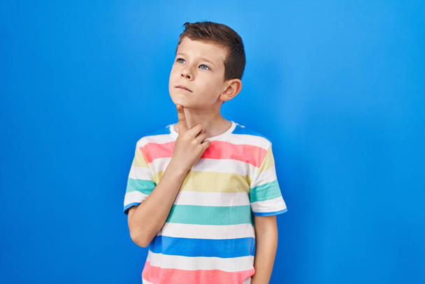 Young caucasian kid standing over blue background thinking concentrated about doubt with finger on chin and looking up wondering  - Foto, afbeelding