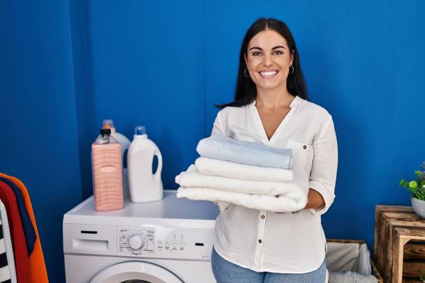 Young beautiful hispanic woman smiling confident holding folded towels at laundry room - Photo, image