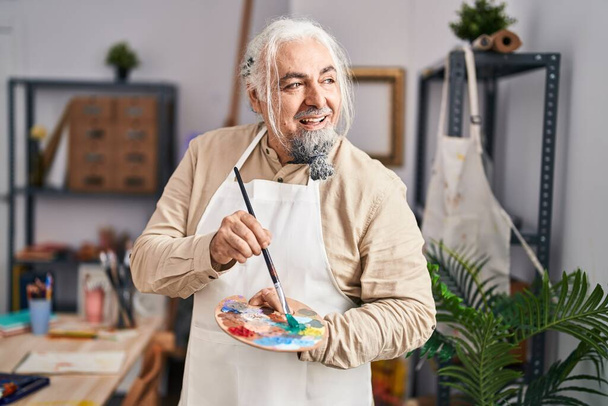 Middle age grey-haired man artist smiling confident holding paintbrush and palette at art studio - Photo, Image