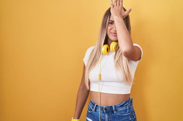 Young blonde woman standing over yellow background wearing headphones surprised with hand on head for mistake, remember error. forgot, bad memory concept.  - Фото, изображение