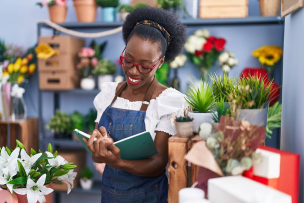 Африканская американка флористка, уверенно улыбающаяся, пишущая на блокноте в цветочном магазине - Фото, изображение