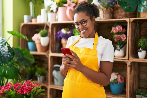 Florista mujer afroamericana sonriendo confiada usando teléfono inteligente en la tienda de flores - Foto, imagen