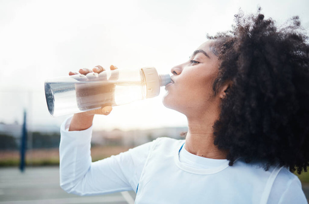 Black woman, sports and drinking water at court during training, workout and sport exercise outdoors. Fitness, thirsty and girl with bottle for wellness, hydration and recovery during practice. - Fotó, kép