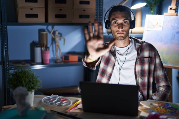 Mladý hispánec, který sedí v ateliéru s laptopem pozdě v noci, přestane zpívat dlaní. varovný výraz s negativním a vážným gestem na tváři.  - Fotografie, Obrázek