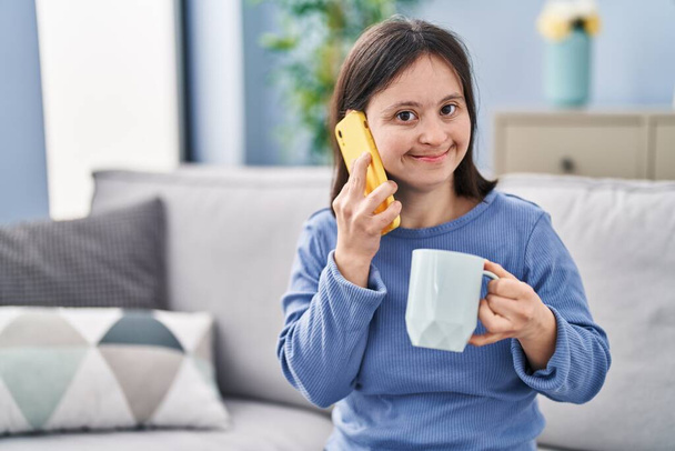 Mujer joven con síndrome de Down beber café y hablar en el teléfono inteligente en casa - Foto, Imagen