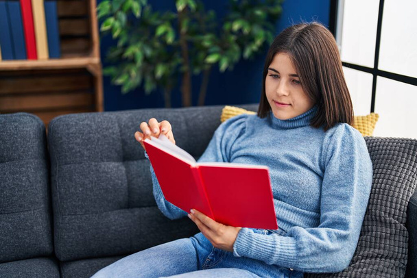 Νεαρή όμορφη ισπανόφωνη γυναίκα διαβάζει το βιβλίο κάθεται στον καναπέ στο σπίτι - Φωτογραφία, εικόνα