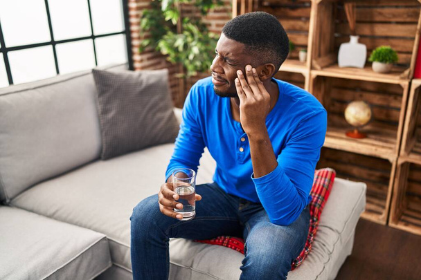 Jonge Afro-Amerikaanse man met hoofdpijn met glas water in huis - Foto, afbeelding
