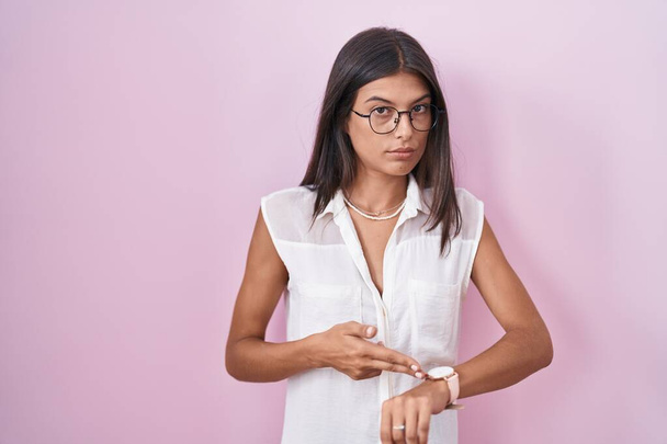 Brunette young woman standing over pink background wearing glasses in hurry pointing to watch time, impatience, upset and angry for deadline delay  - Фото, зображення
