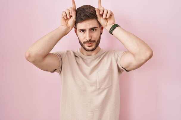 Spanyol férfi szakállal áll a rózsaszín háttér csinál vicces gesztus az ujját a feje fölött, mint a bika szarv  - Fotó, kép