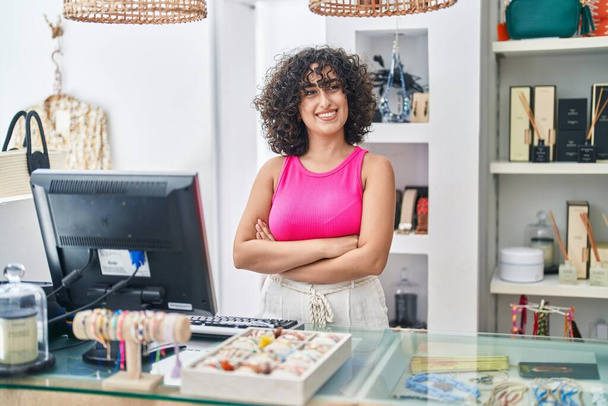 Giovane donna mediorientale commessa sorridente fiducioso in piedi con le braccia incrociate gesto al negozio di abbigliamento - Foto, immagini