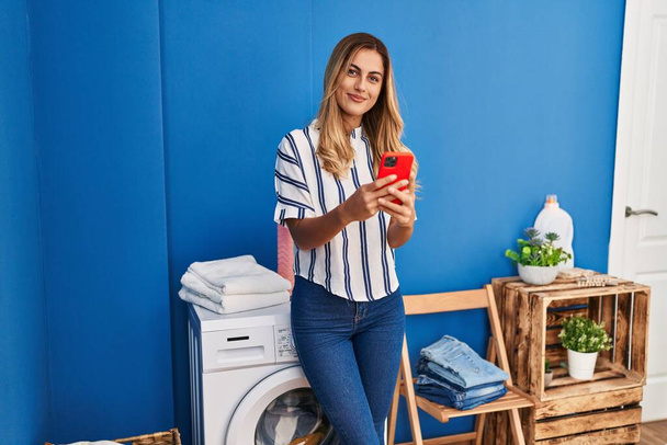 Jovem loira usando smartphone esperando máquina de lavar roupa na lavanderia - Foto, Imagem